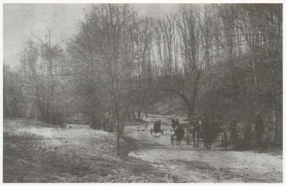 Vintage photo of horse-drawn carriages crossing a river in a wooded area.